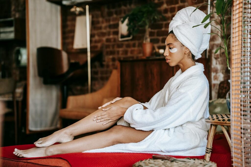 Side view of young female in white robe and white towel on head relaxing on comfortable bed and applying cream on leg